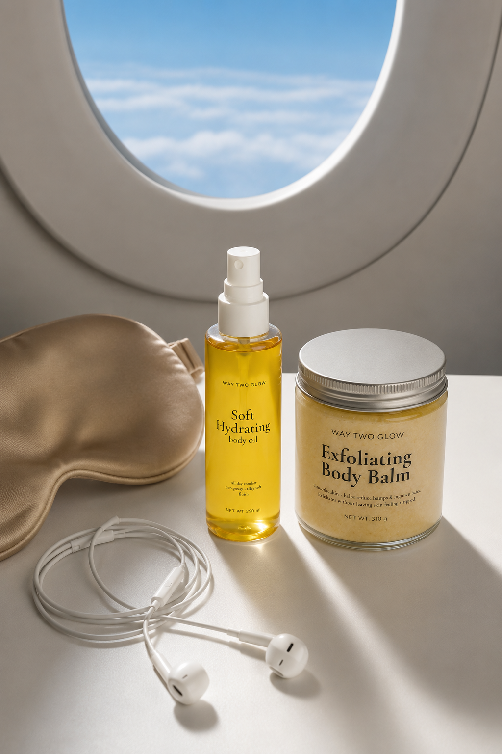 A travel-themed skincare set on a white surface with an airplane window background. The set includes a gold satin sleep mask, a bottle of yellow 'Soft Hydrating Body Mist', a jar of 'Exfoliating Body Balm' with a silver lid, and white wired earbuds.